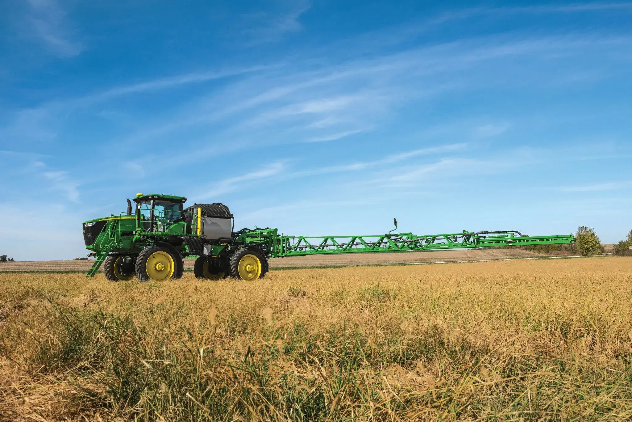 A large green John Deere agricultural sprayer vehicle is positioned in a field of tall, dry grass under a clear blue sky. The vehicle has yellow wheels and an extended arm for spraying crops, showcasing advanced farming machinery in a rural setting.